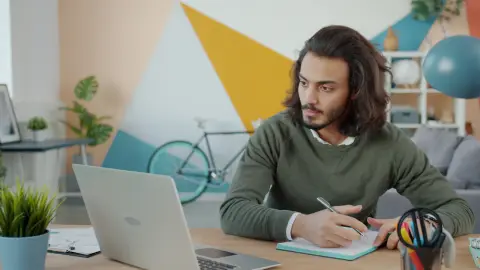 A man taking notes beside an open laptop at a desk