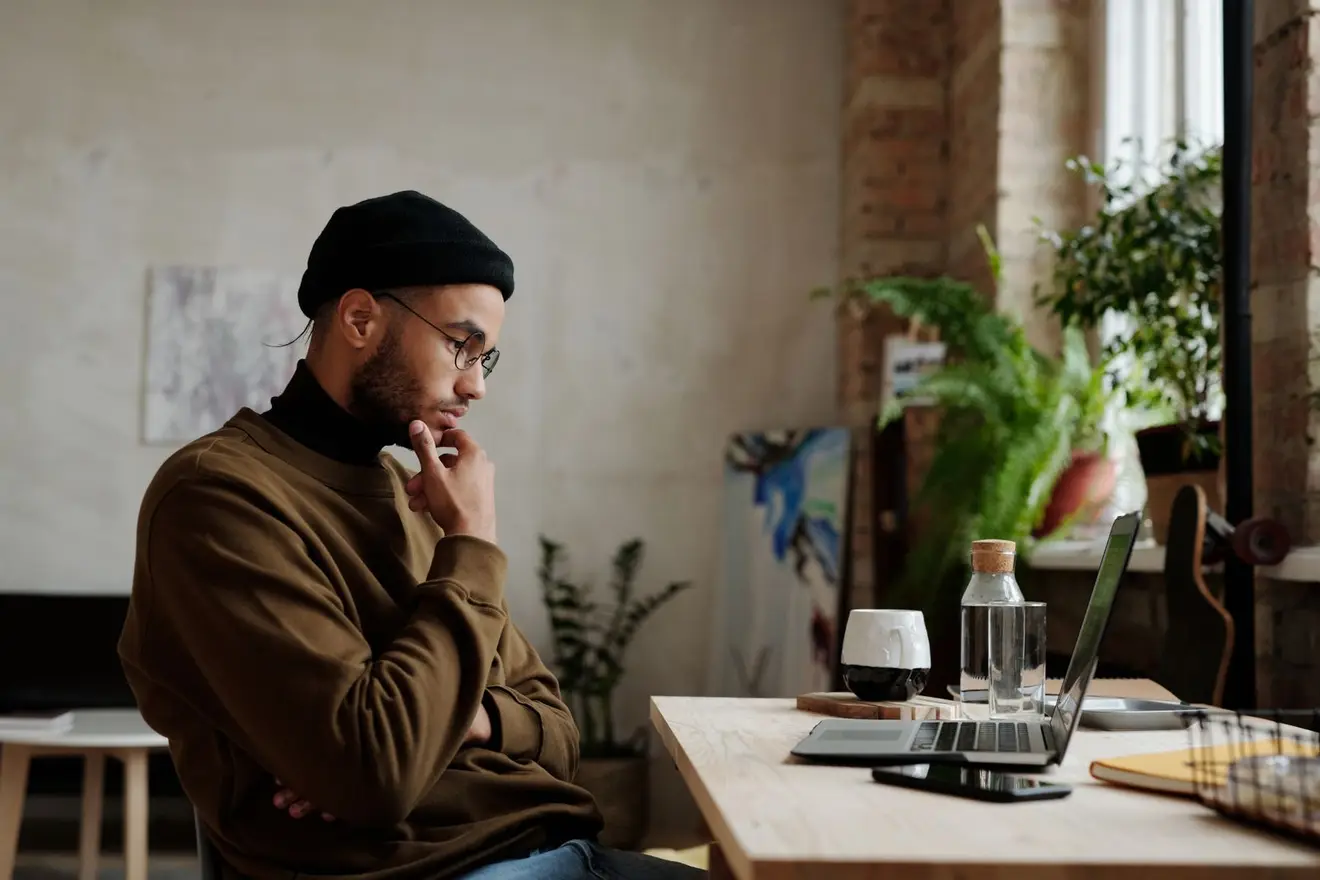 A man sitting at a table, looking thoughtfully at his laptop