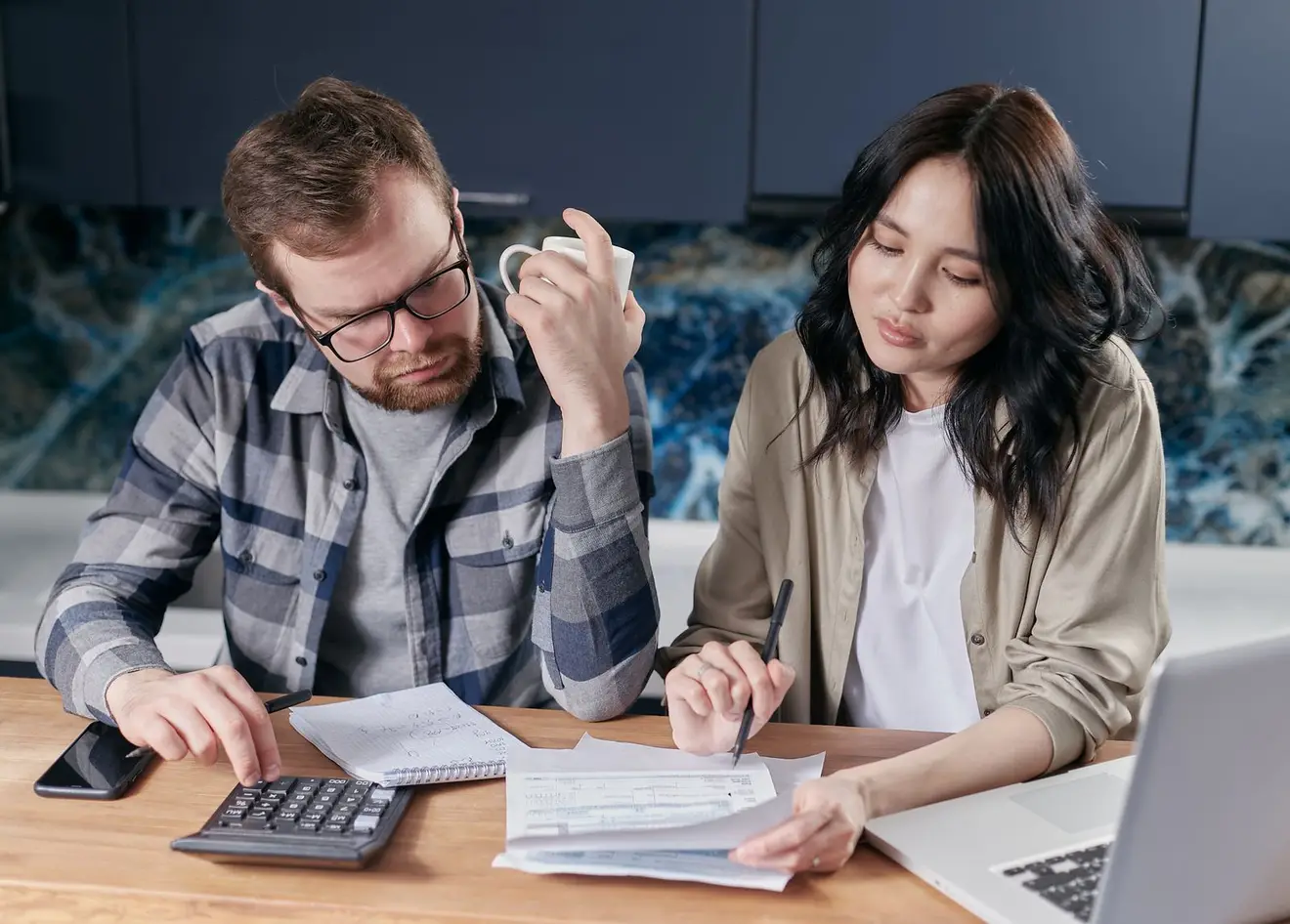 A couple reviewing household bills with a calculator and laptop at a kitchen table