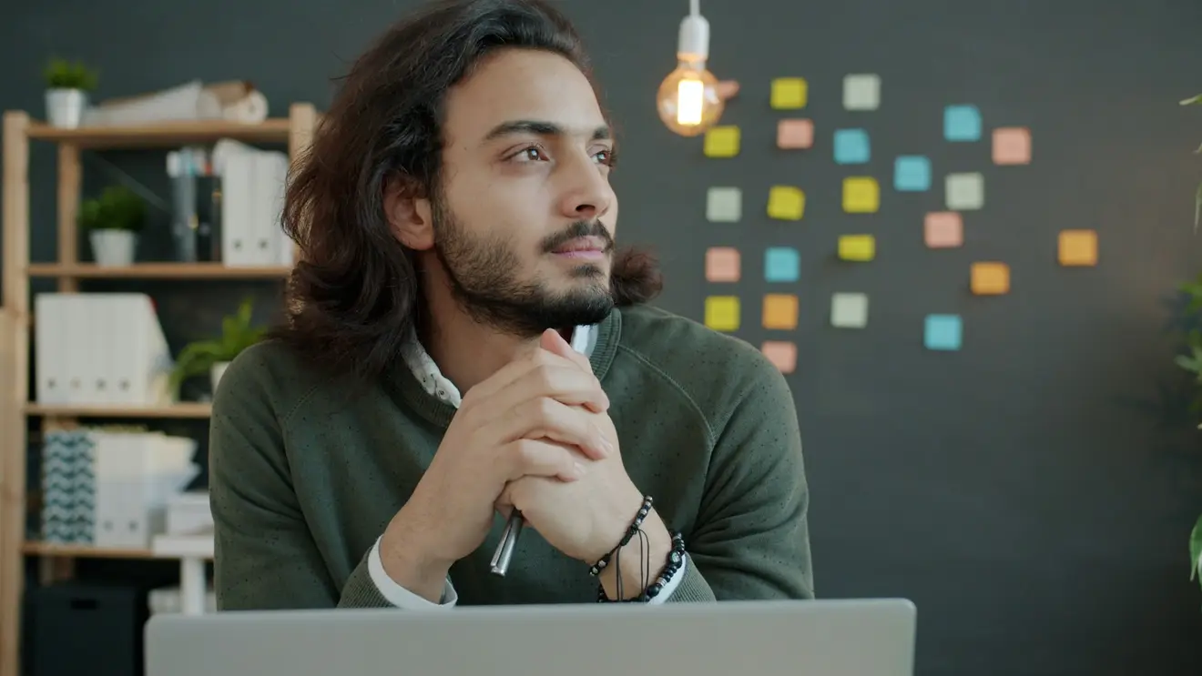 A man pauses to think with a pen in his hands and a laptop in front of him.