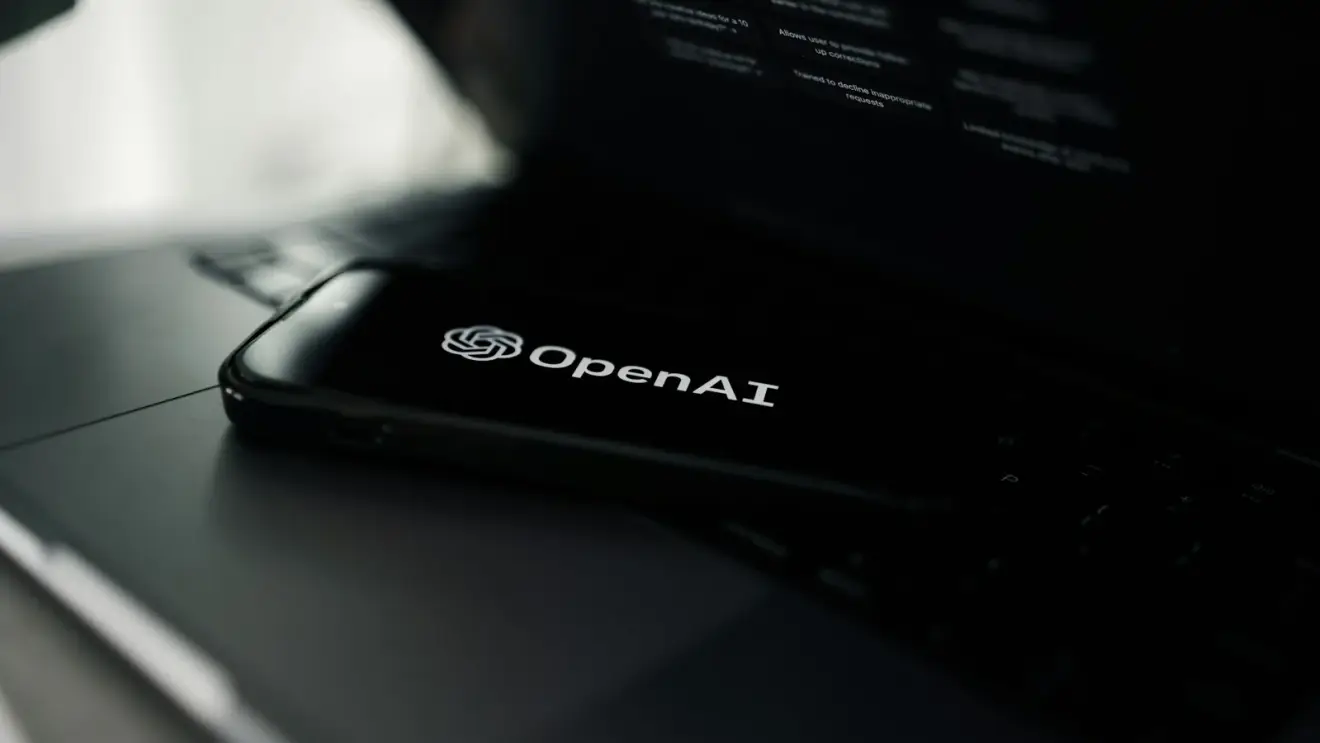 OpenAI logo displayed on a phone resting on a laptop showing ChatGPT