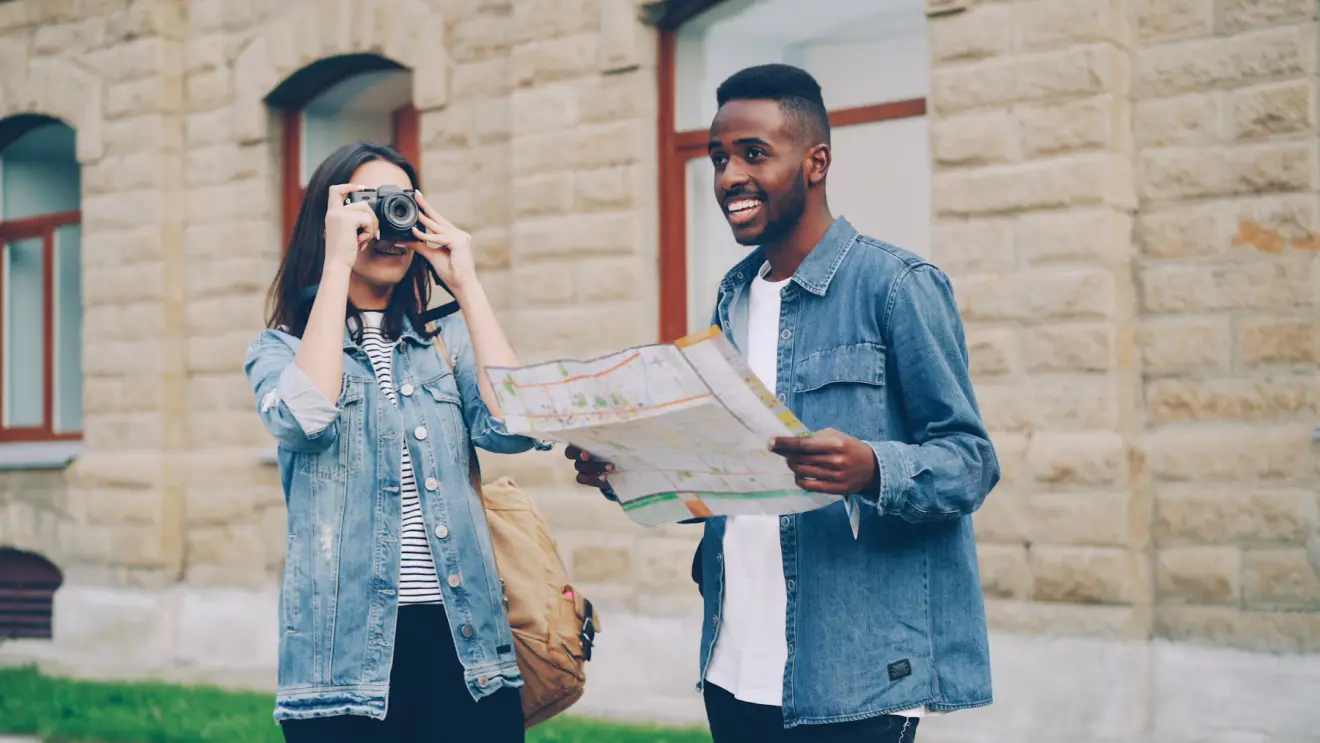 Two travelers outdoors, one holding a paper map and the other taking a photograph.